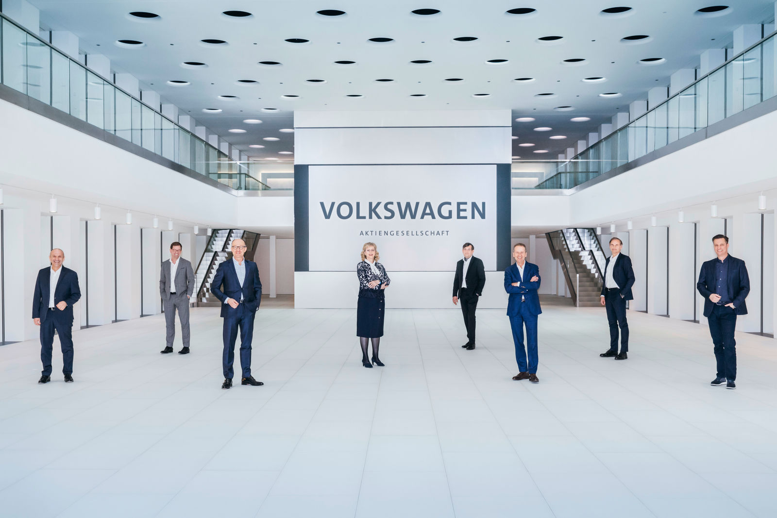 A group of seven people in formal attire standing in a modern, bright space in front of a large Volkswagen logo.
