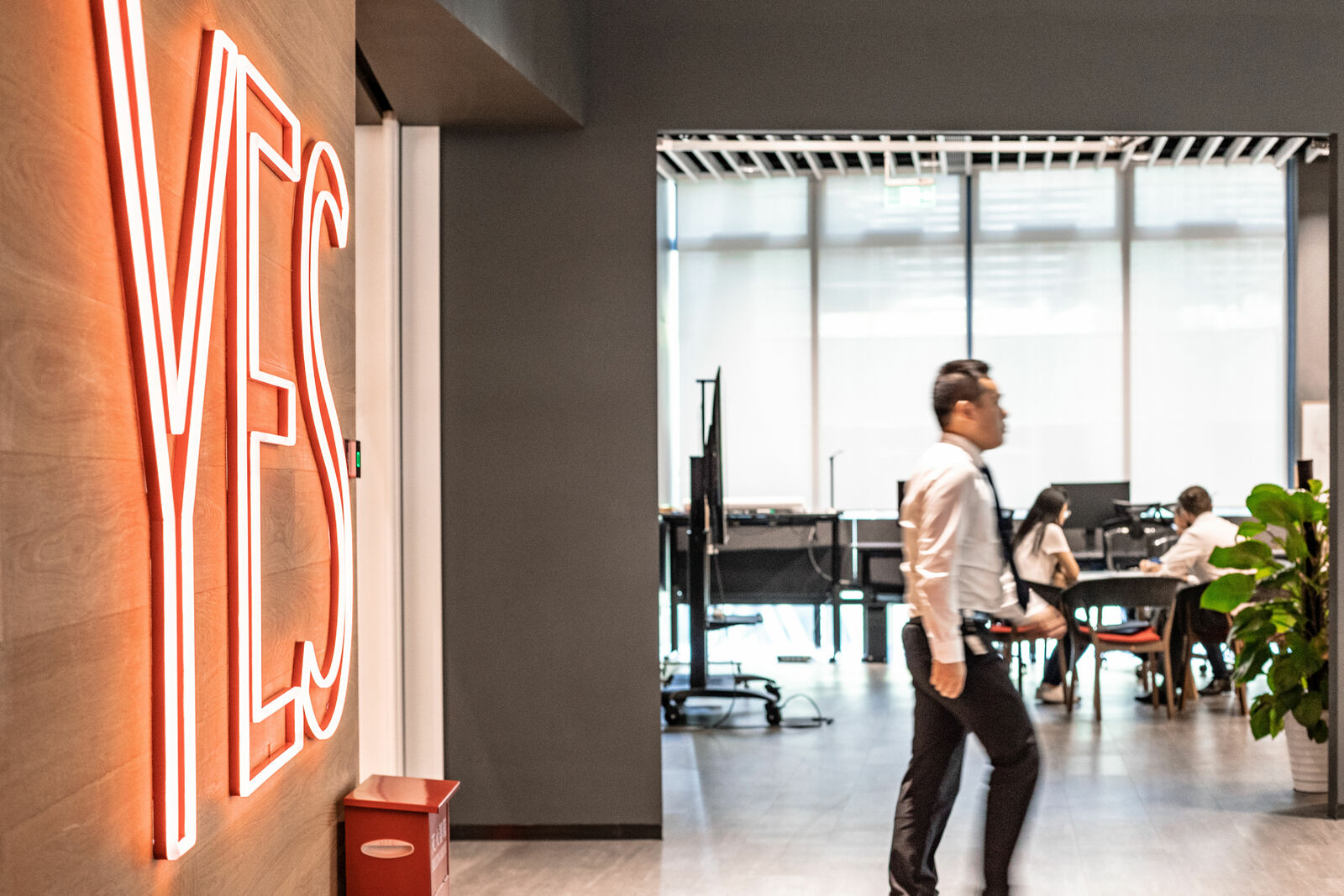Ein modernes Büro mit einem roten Neon-Schriftzug „YES“ an der Wand. Im Vordergrund geht ein Mann in Businesskleidung vorbei, während im Hintergrund mehrere Personen an einem Besprechungstisch arbeiten. Große Fenster sorgen für viel Tageslicht.