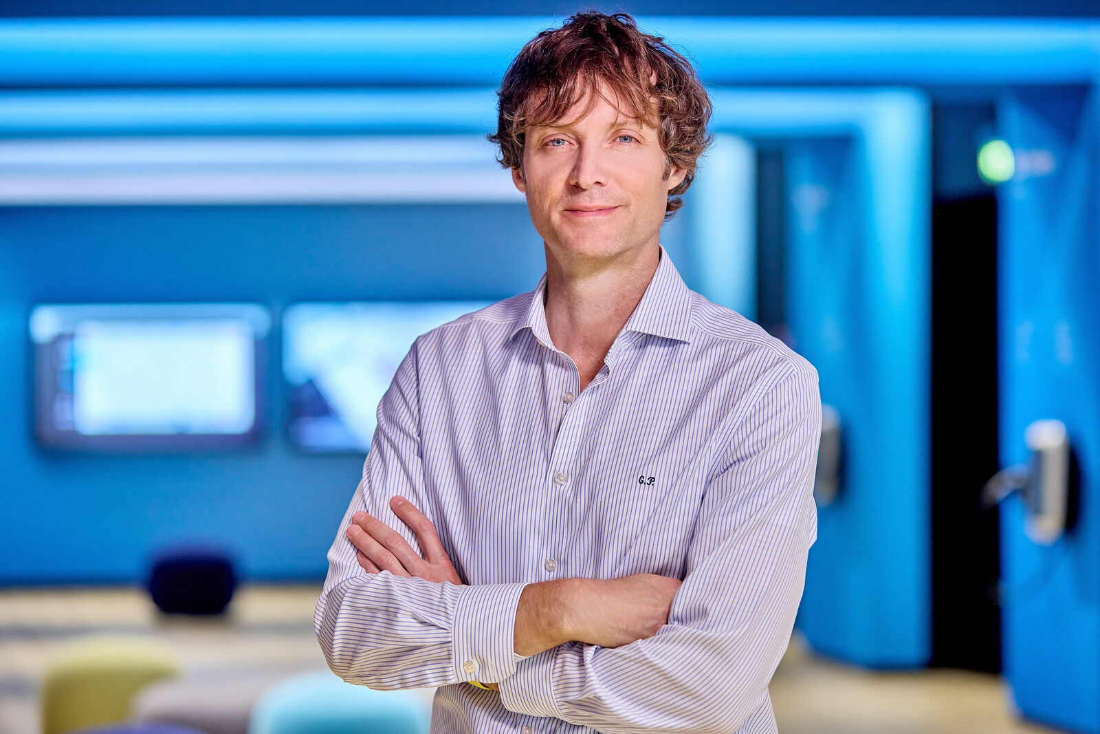 A man with curly hair and a light-colored shirt stands with crossed arms in a modern office space with blue lighting. Screens and seating areas are visible in the background.