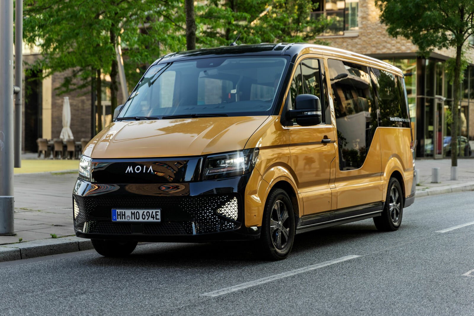 A gold-black MOIA electric shuttle van is parked on a city street. The vehicle features a modern design with a curved front, a black grille, and LED headlights. The large side door allows for easy entry. In the background, modern buildings, trees, and a pedestrian area are visible. The license plate reads "HH MO 694E," indicating registration in Hamburg.