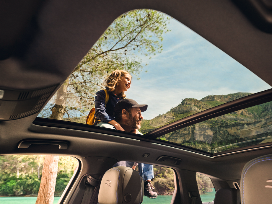 A man and a child stand outside a car, looking at mountains, captured from inside the car through the open panoramic roof.