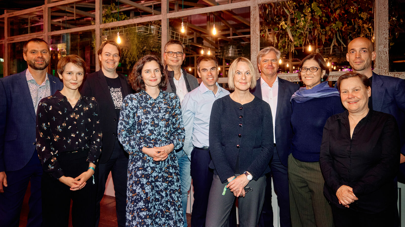 A group of eleven people poses indoors with plants and decorative lights in the background. Everyone is smiling and standing in a relaxed formation.