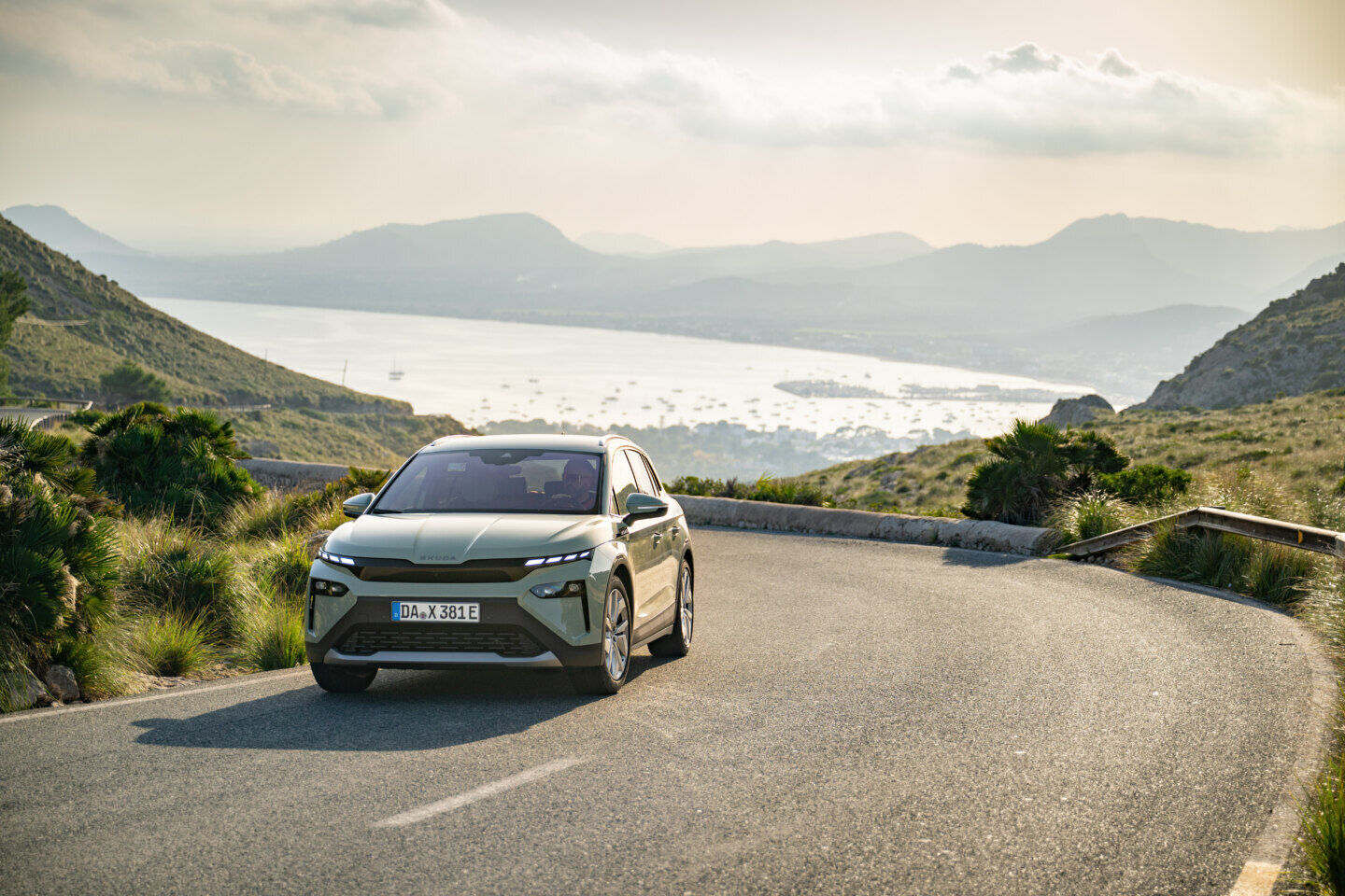 Škoda electric vehicle driving on a winding mountain road overlooking a coastal landscape.